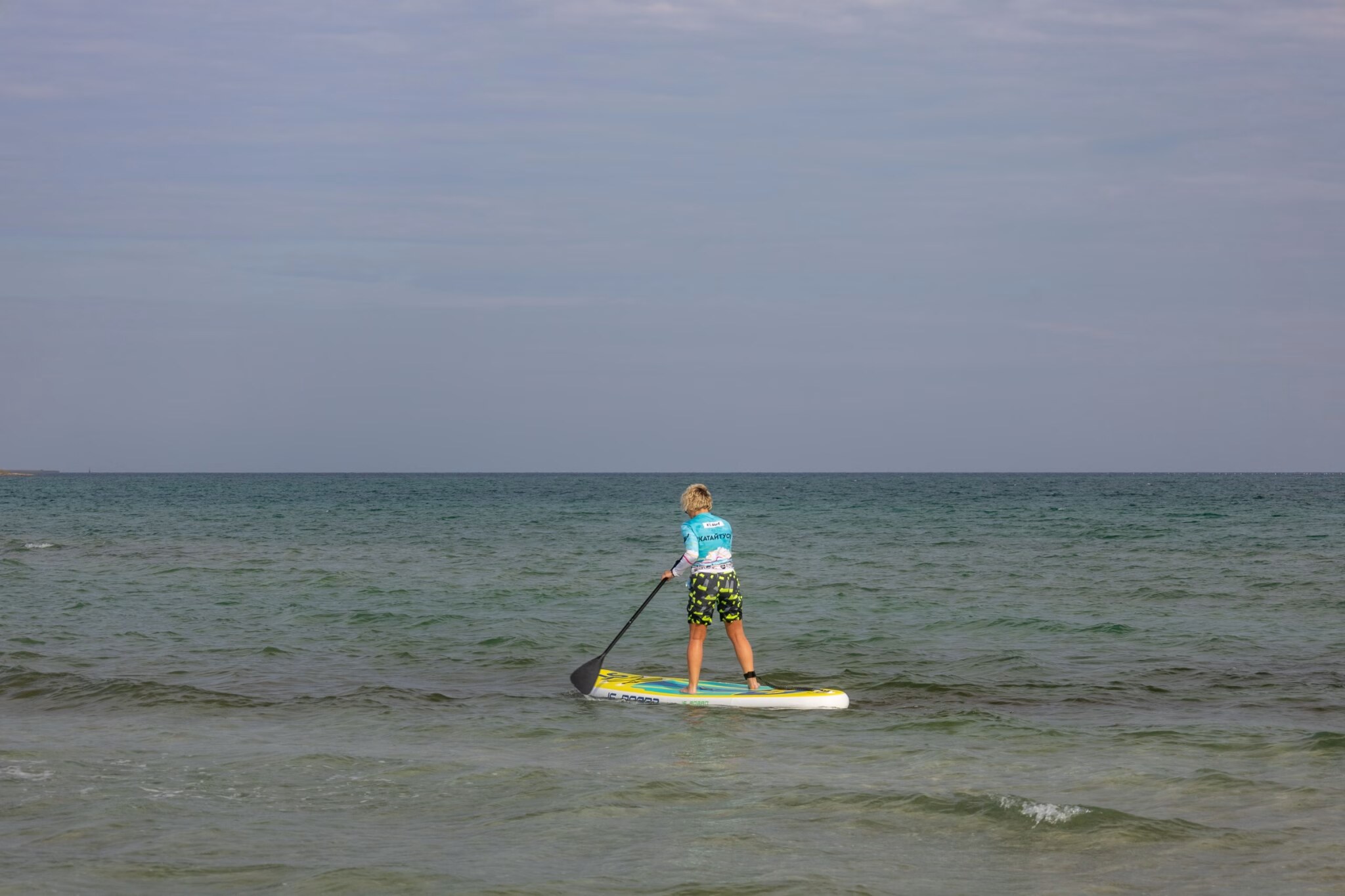 Stand-up paddleboarder mid-stroke on calm open water, torso rotated and top arm extended at the catch