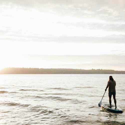 Paddle boarder silhouette on calm water at sunset, paddle in hand, illustrating the sustained grip and arm positioning that loads peripheral nerves during long sessions