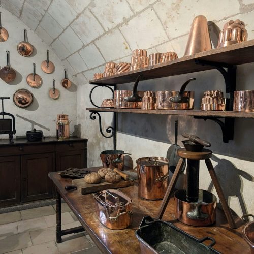 A historic kitchen lined with polished copper pots, pans, and fluted copper molds on wooden shelves and whitewashed stone walls
