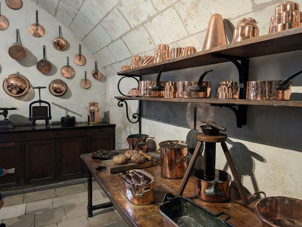 A historic kitchen lined with polished copper pots, pans, and fluted copper molds on wooden shelves and whitewashed stone walls