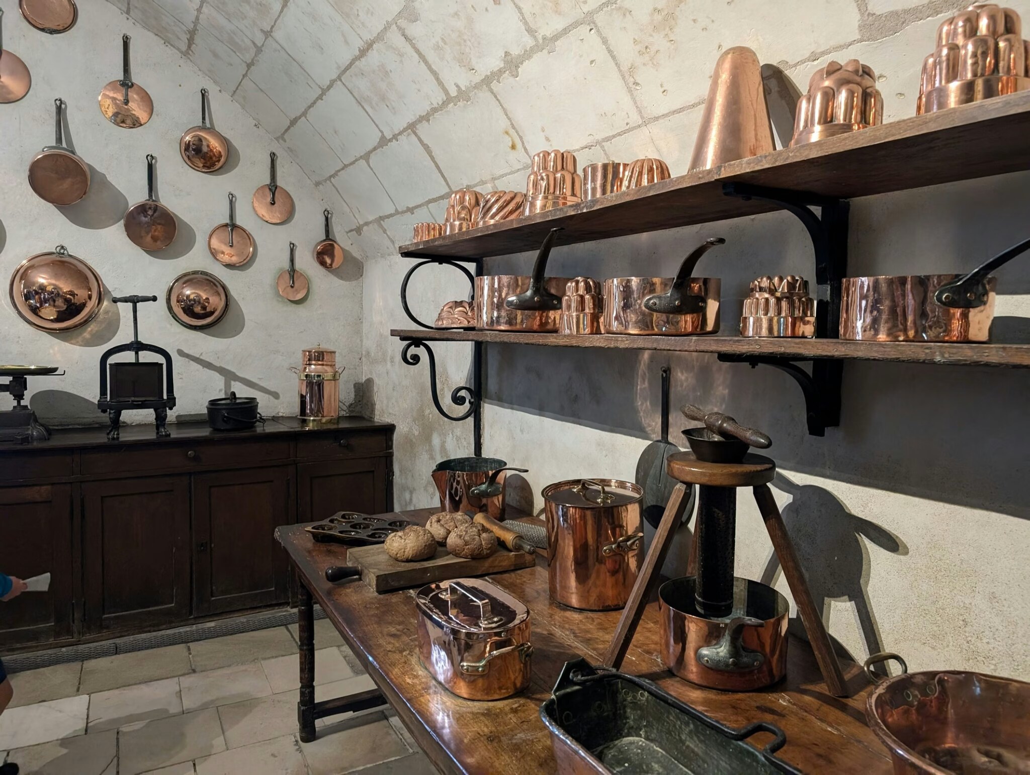 A historic kitchen lined with polished copper pots, pans, and fluted copper molds on wooden shelves and whitewashed stone walls