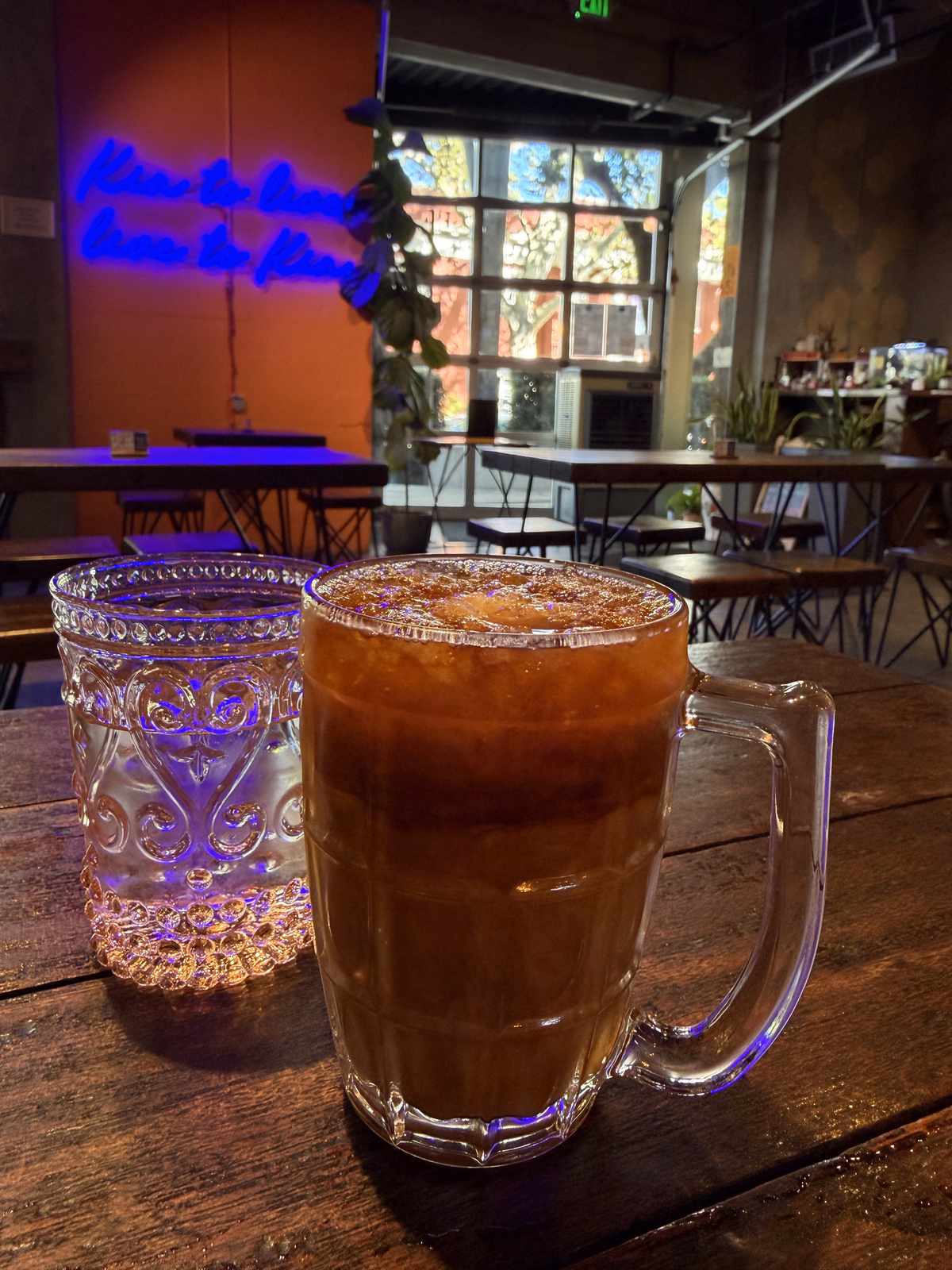 Thai iced tea in a glass mug on a wooden table at kin thai street eatery, with blue neon script glowing on the wall behind