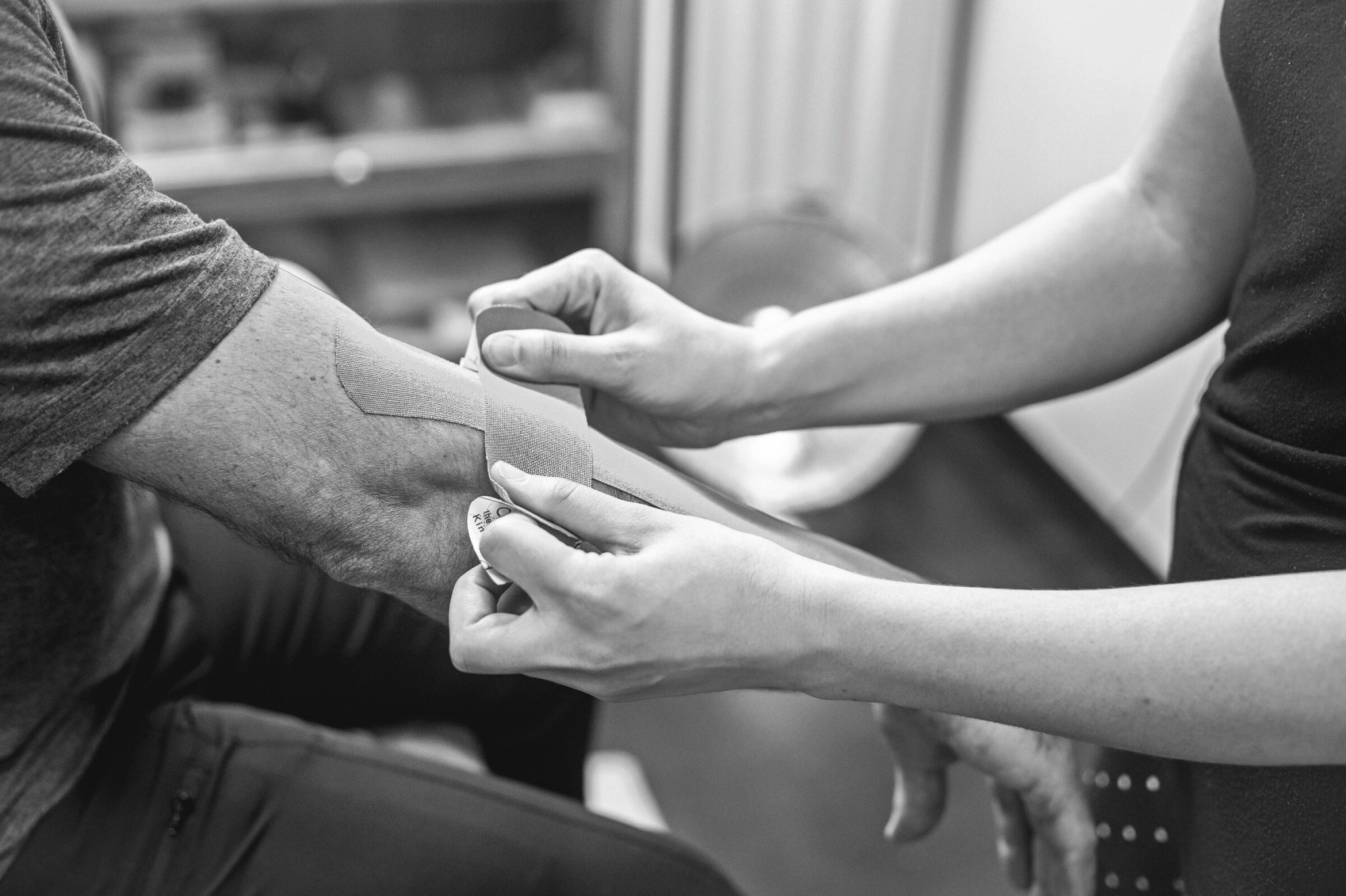 Black and white close-up of a clinician laying a strip of kinesiology tape along a patient's forearm toward the elbow