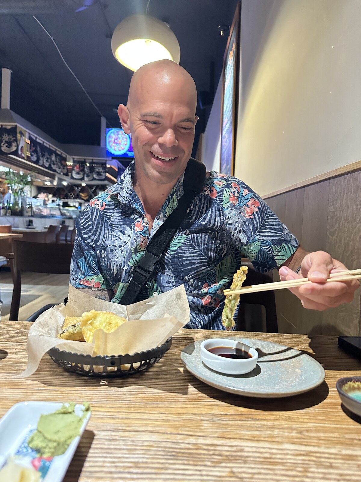 Michael kahn at a restaurant table picking up tempura with chopsticks in his left hand, a sling strap across his chest