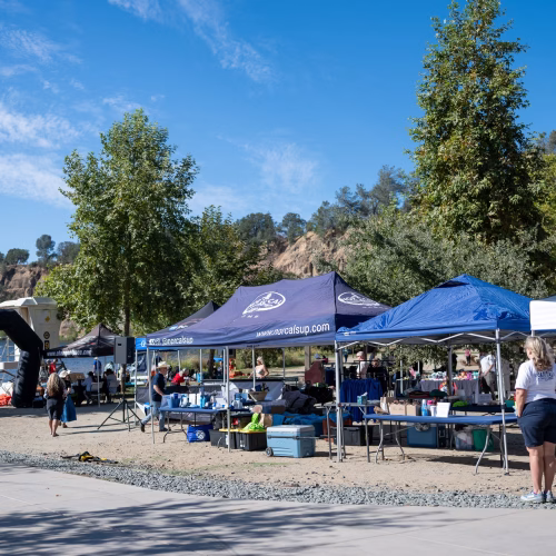 Inflatable finish arch on the sandy beach at black miner's bar, with nor cal sup and visit folsom tents and vendor tables set up behind it beside lake natoma