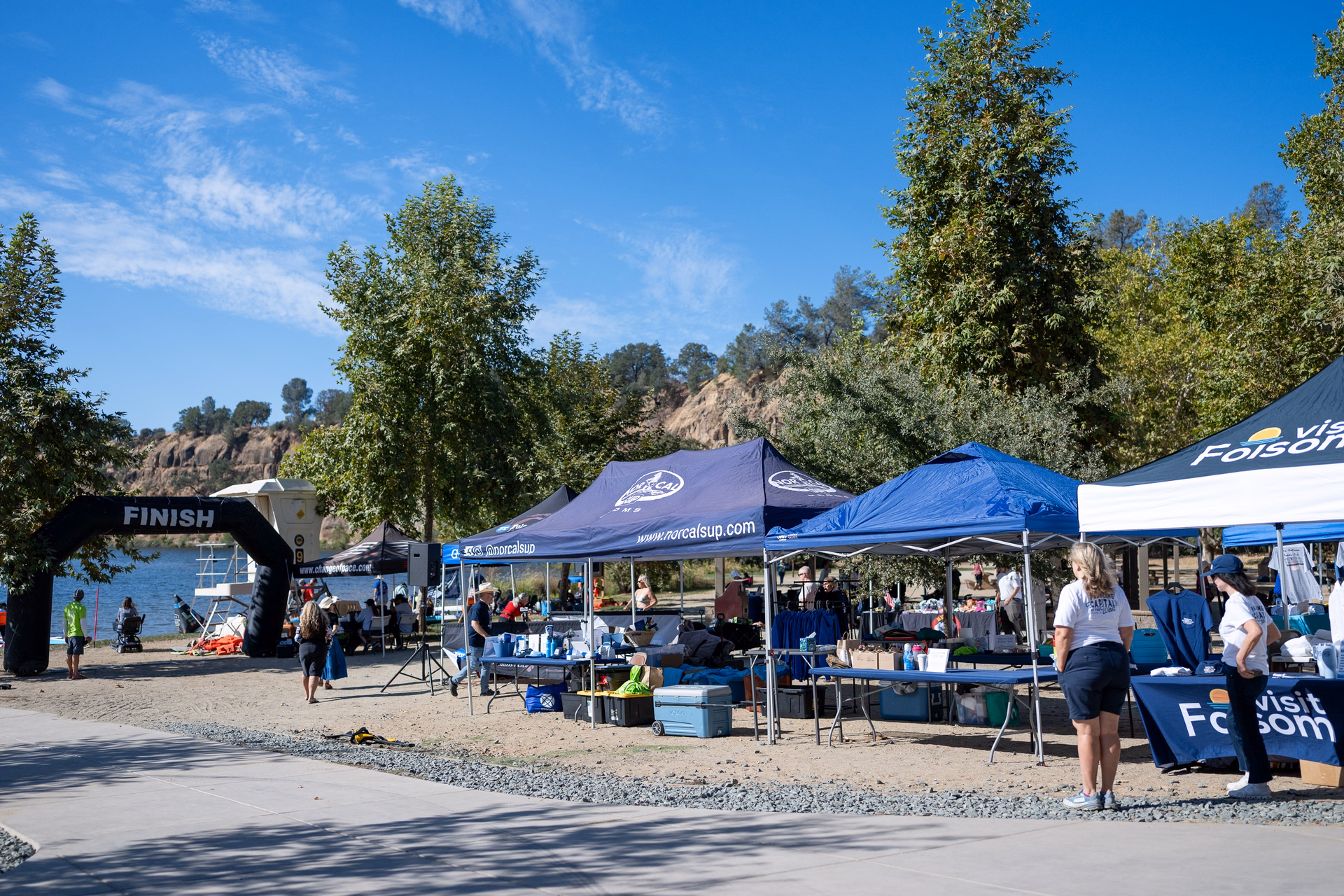 Inflatable finish arch on the sandy beach at black miner's bar, with nor cal sup and visit folsom tents and vendor tables set up behind it beside lake natoma