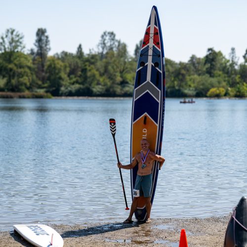 Paddler standing beside his 14-foot venture wild sierra pro inflatable board stood on end, wearing race medals and bib 36, at lake natoma