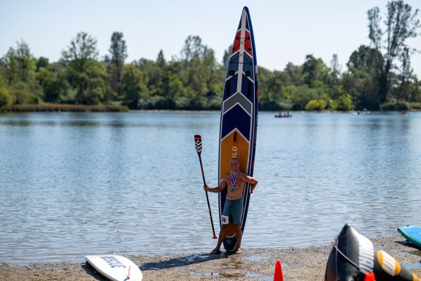 Paddler standing beside his 14-foot venture wild sierra pro inflatable board stood on end, wearing race medals and bib 36, at lake natoma