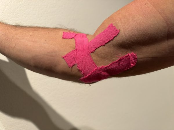 Pink kinesiology tape on a right elbow, strips anchored on the forearm below the crease and running up across the front of the elbow onto the upper arm