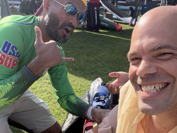 Michael kahn smiling on the grass at usa sup nationals in gig harbor with his right arm wrapped and a sling tied at his chest, while carl, a nurse practitioner on the event medical team, holds the wrap and gives a shaka