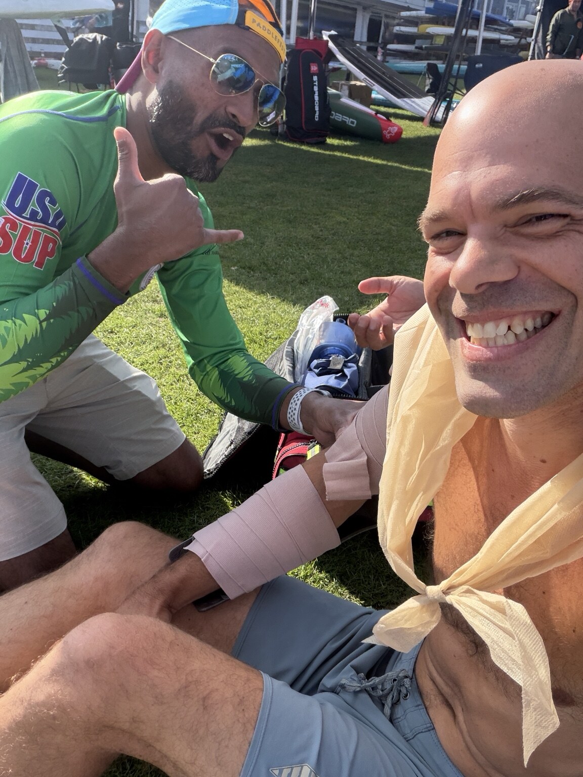 Michael kahn sitting on the grass at usa sup nationals in gig harbor with his right arm wrapped in an elastic bandage and a sling tied at his chest, while carl, a nurse practitioner on the event medical team, holds the wrap and gives a shaka