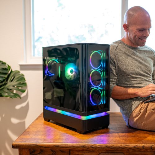 Michael kahn sits on an oak desk typing on a keyboard beside the finished gaming pc, its fan rings and base light band lit behind dark glass, with a monstera plant at left
