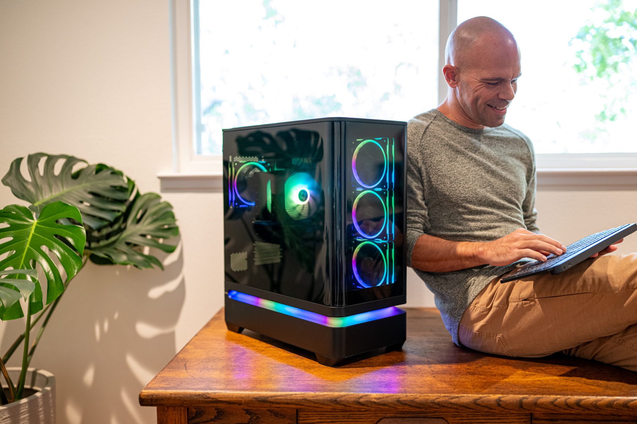 Michael kahn sits on an oak desk typing on a keyboard beside the finished gaming pc, its fan rings and base light band lit behind dark glass, with a monstera plant at left