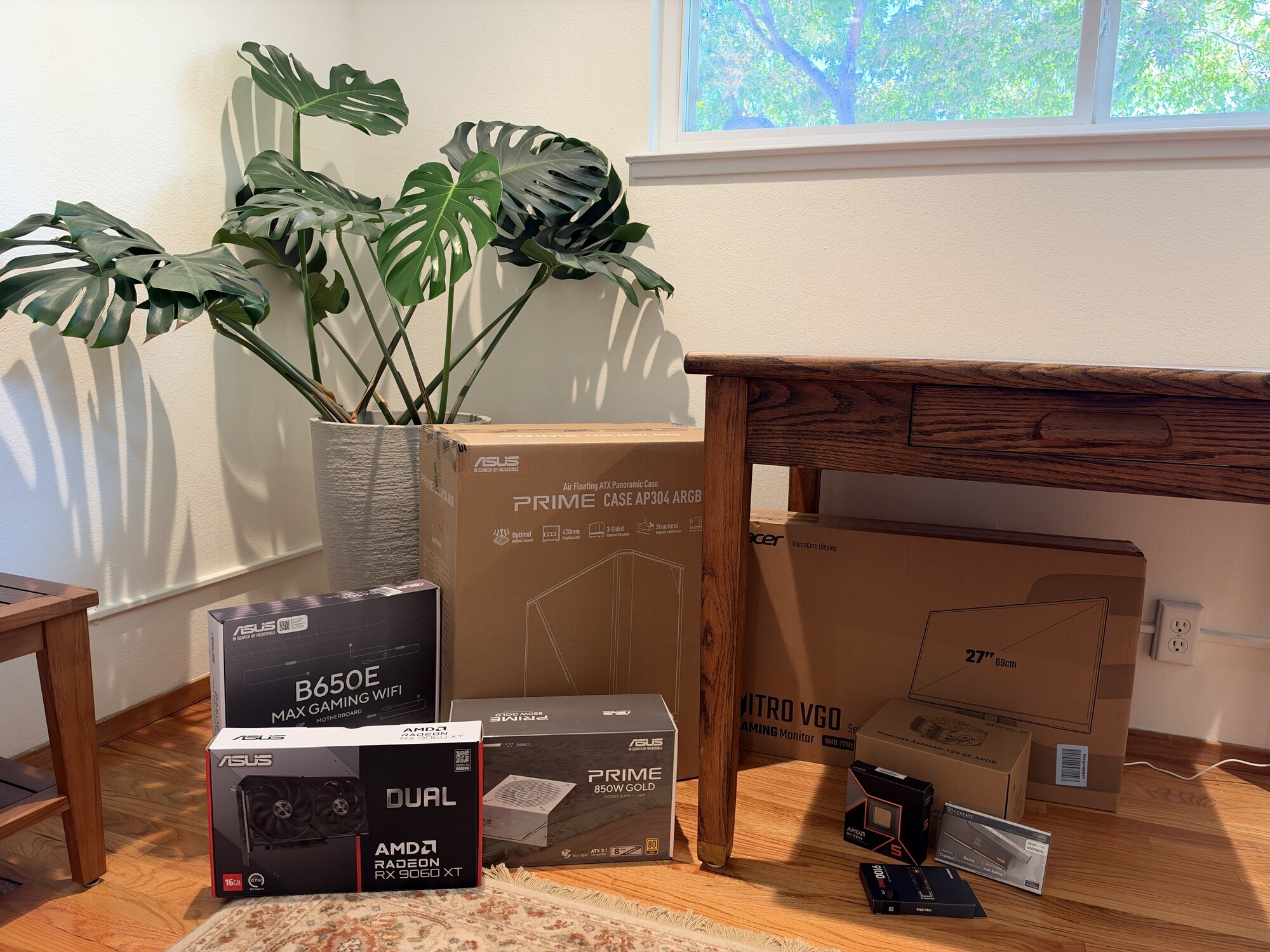 Boxed pc parts on a hardwood floor beside a monstera plant, including an asus prime ap304 argb case, motherboard, graphics card, power supply and acer monitor