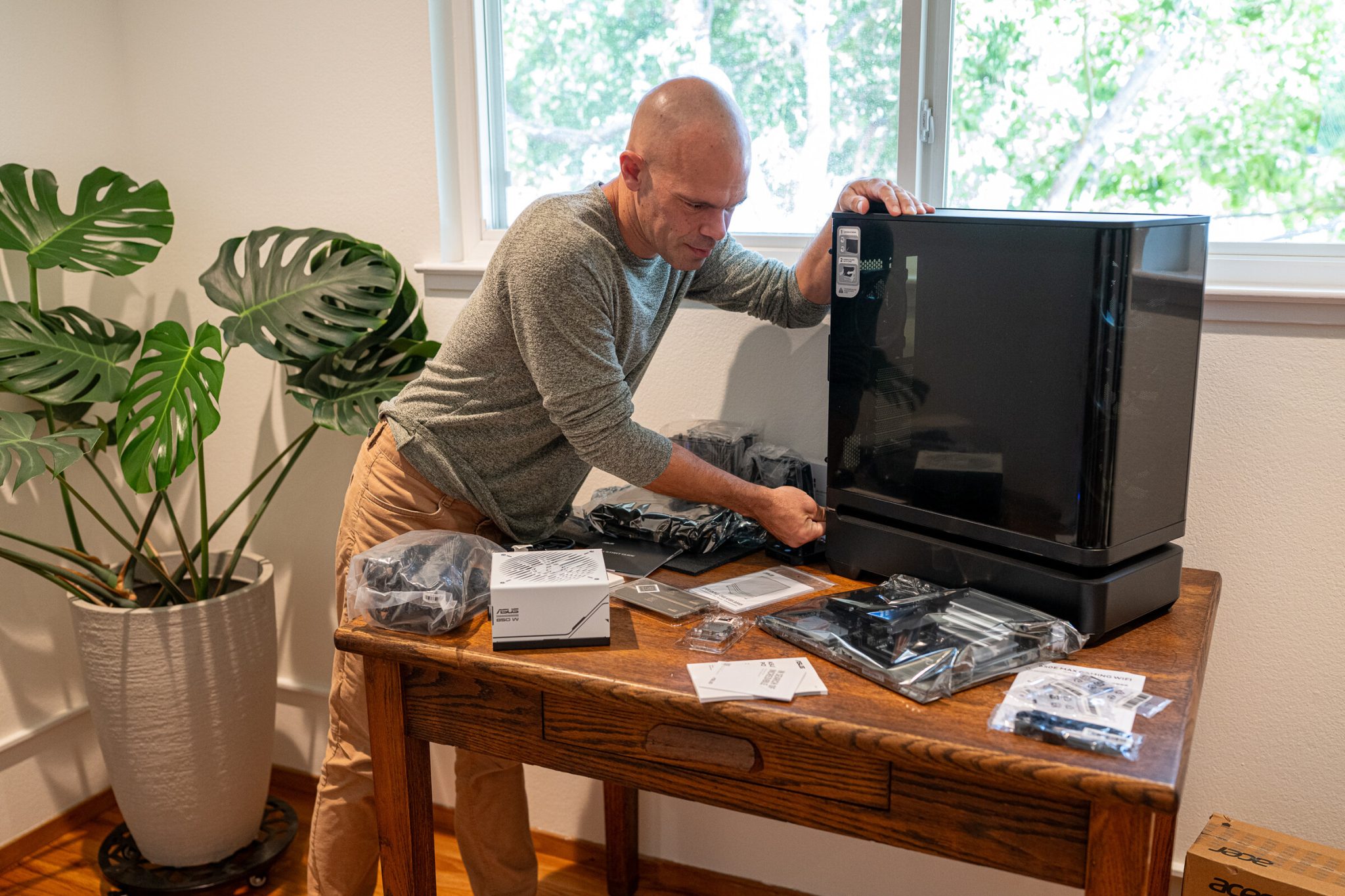 Michael kahn stands at an oak desk beside a black asus prime ap304 case, with a white power supply, a bagged motherboard and bags of cables laid out in front of him