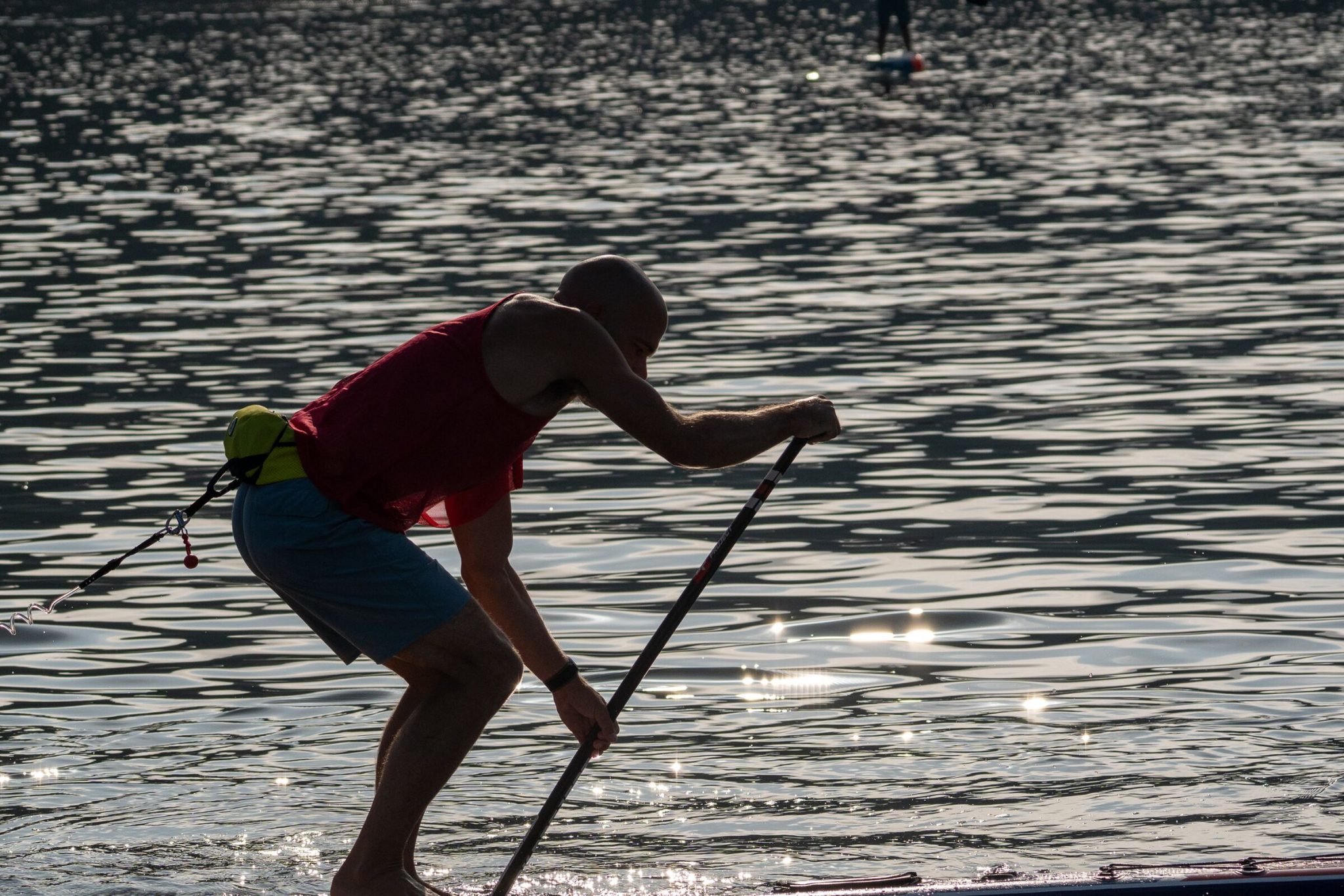 Silhouette of a stand-up paddler bent into a stroke with both hands on the paddle shaft against sunlit water at the 2026 usa sup national championships