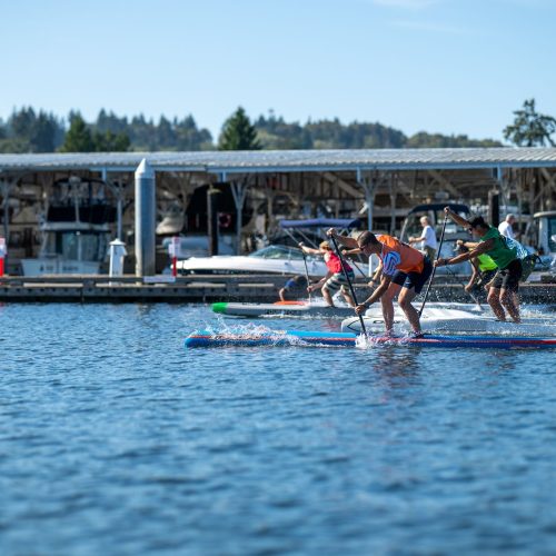 Three stand-up paddlers bent into their strokes on race boards, throwing spray as they sprint past the gig harbor docks at the 2026 usa sup national championships