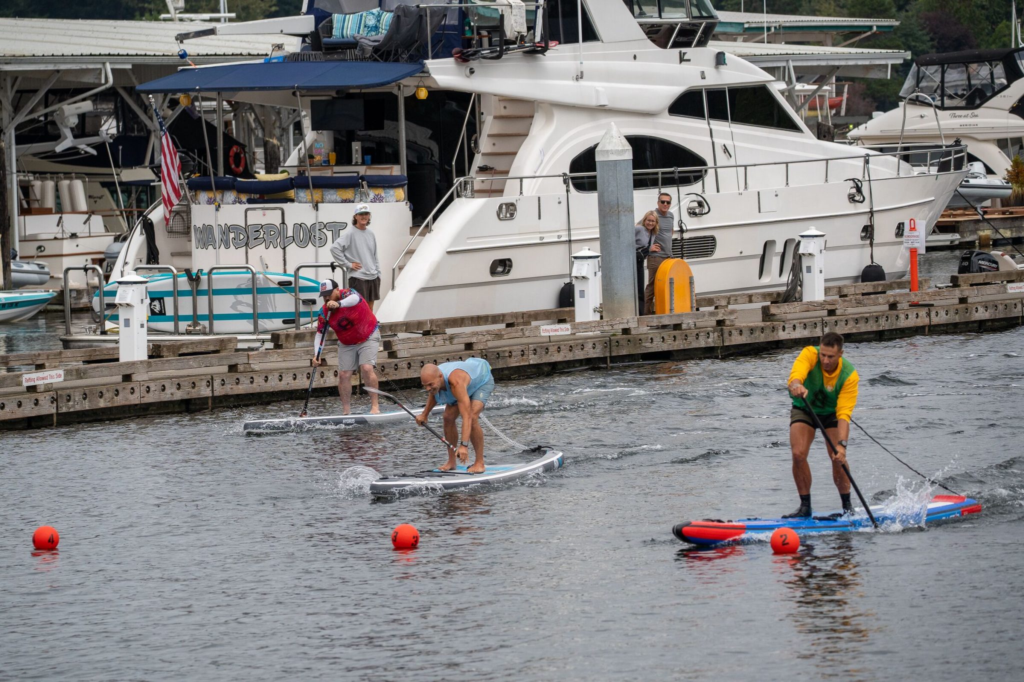 Three paddlers on inflatable race boards sprinting past orange lane buoys beside the docks in gig harbor, one in a blue tank top